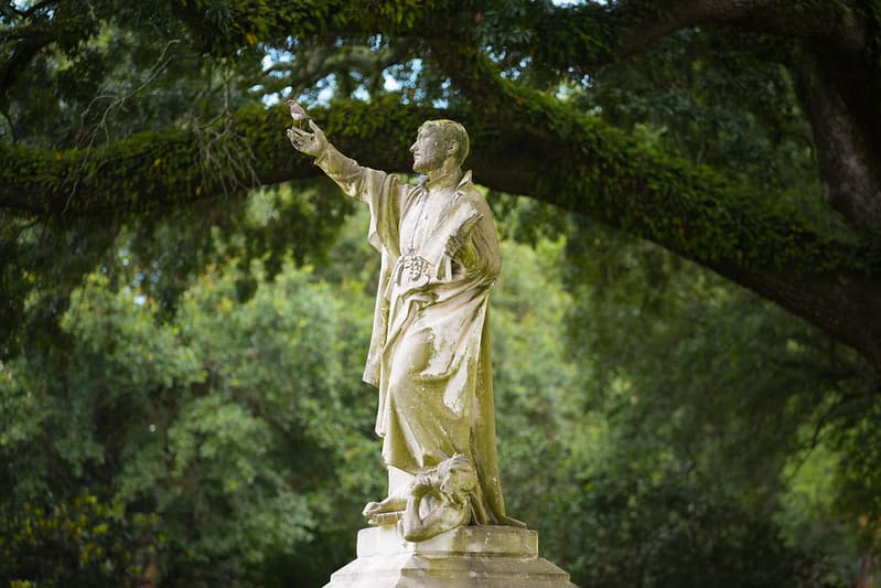 Campus statue of a Jesuit reaching toward a bird beneath the live oaks